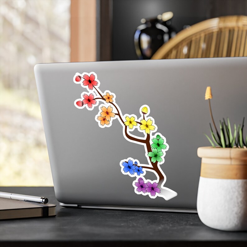 Pu&ograve; includere: Un adesivo di un ramo di ciliegio in fiore arcobaleno con uno stelo marrone e foglie verdi. L'adesivo &egrave; su un laptop argentato.