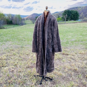 May include: A long, brown, fuzzy coat with a shawl collar. The coat is on a mannequin and is standing in a field.
