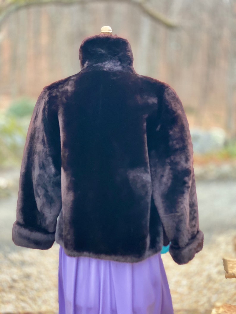 May include: A brown faux fur jacket with a high collar and long sleeves. The jacket is on a mannequin and the background is blurred.