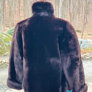 May include: A brown faux fur jacket with a high collar and long sleeves. The jacket is on a mannequin and the background is a blurry image of trees.