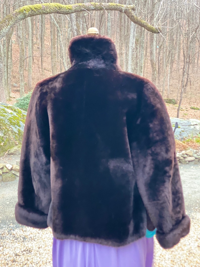 May include: A brown faux fur jacket with a high collar and long sleeves. The jacket is on a mannequin and the background is a blurry image of trees.