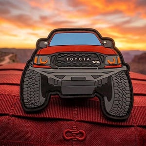 May include: A red and black patch featuring a stylized Toyota truck with large tires. The patch has the word "TOYOTA" across the front and "TACO." below. The background is a sunset over a desert landscape.