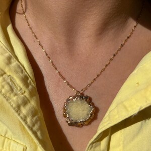 Close-up of a raw yellow druzy crystal pendant necklace with an irregular floral-like shape and gold-toned electroplated edges. The pendant hangs from a delicate twisted gold chain, resting against a woman's neck dressed in a light yellow shirt. The natural texture of the stone sparkles under sunlight, adding earthy elegance and bohemian charm.