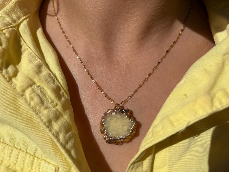 Close-up of a raw yellow druzy crystal pendant necklace with an irregular floral-like shape and gold-toned electroplated edges. The pendant hangs from a delicate twisted gold chain, resting against a woman's neck dressed in a light yellow shirt. The natural texture of the stone sparkles under sunlight, adding earthy elegance and bohemian charm.