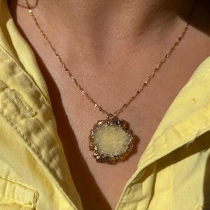 Close-up of a raw yellow druzy crystal pendant necklace with an irregular floral-like shape and gold-toned electroplated edges. The pendant hangs from a delicate twisted gold chain, resting against a woman's neck dressed in a light yellow shirt. The natural texture of the stone sparkles under sunlight, adding earthy elegance and bohemian charm.