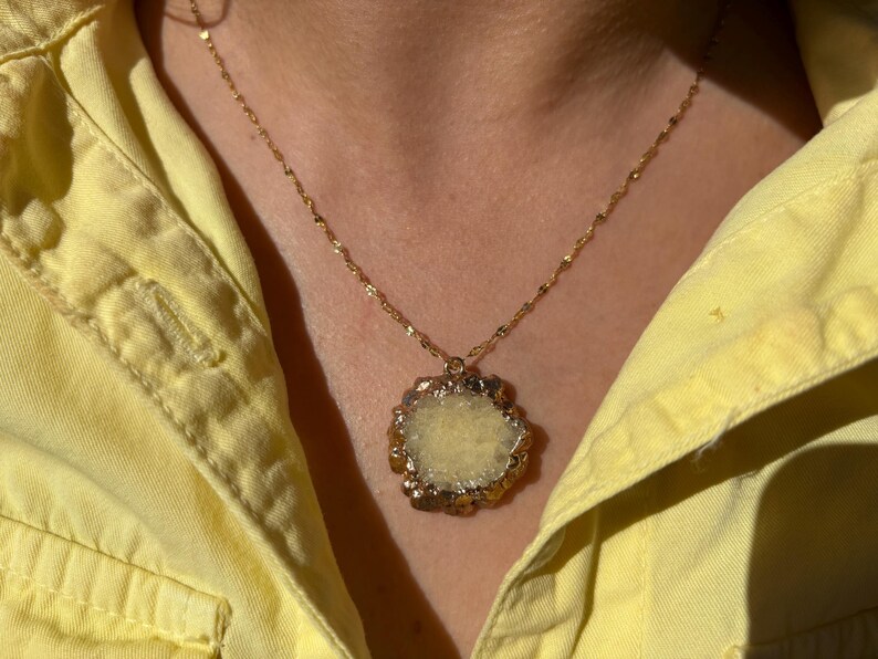 Close-up of a raw yellow druzy crystal pendant necklace with an irregular floral-like shape and gold-toned electroplated edges. The pendant hangs from a delicate twisted gold chain, resting against a woman's neck dressed in a light yellow shirt. The natural texture of the stone sparkles under sunlight, adding earthy elegance and bohemian charm.