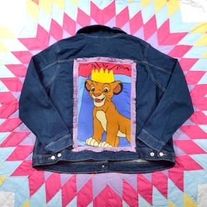 Puede incluir: Una chaqueta vaquera azul con un parche cosido en la espalda. El parche presenta un cachorro de león de dibujos animados con una corona, sobre un fondo morado, azul y amarillo.