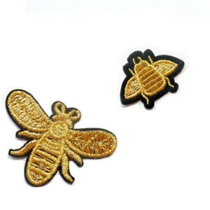 Könnte beinhalten: Zwei goldene gestickte Bienen-Patches. Eine Biene ist größer und detaillierter als die andere. Beide Bienen befinden sich auf einem schwarzen Stoffgrund.