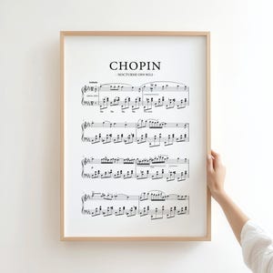 May include: A framed print of musical notation, with the name "CHOPIN" at the top. The sheet music is black on white, in a light brown frame. The print is held by a person.