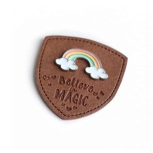 May include: A brown leather patch with a stitched edge in the shape of a shield. The patch has a rainbow enamel pin with white clouds on top of it. The text "Believe in MAGIC" is embossed on the patch.