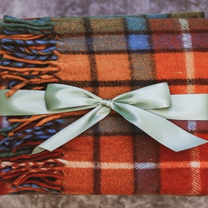 May include: A red, orange, green, and blue plaid throw blanket with a sage green satin ribbon tied in a bow.