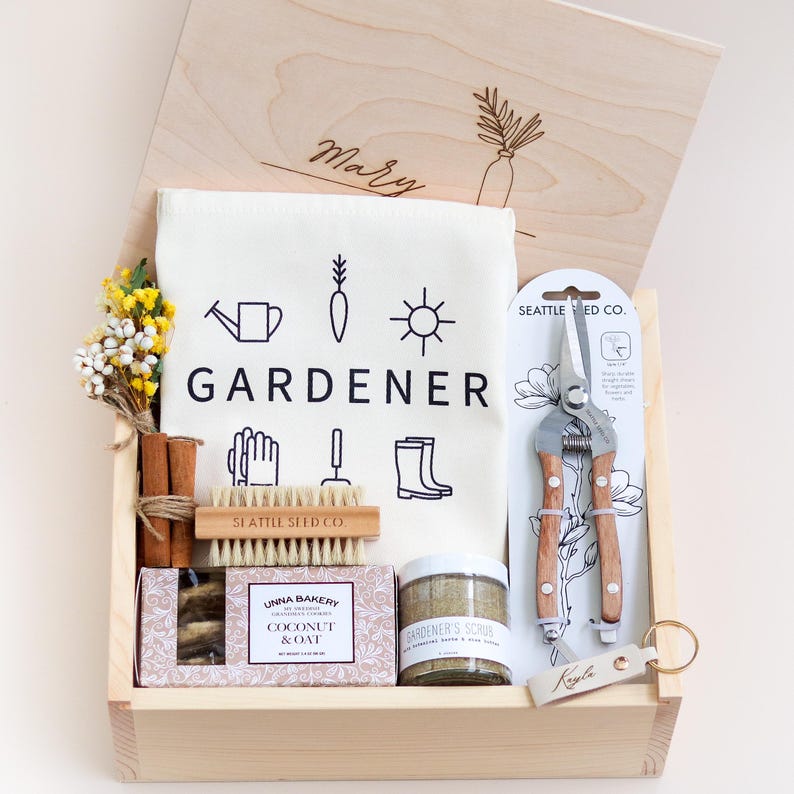 May include: A wooden gift box with the word "GARDENER" printed on a white tea towel. The box contains gardening tools, a bar of soap, a scrub, cinnamon sticks, and a small wooden brush. The box is personalized with the name "Mary".