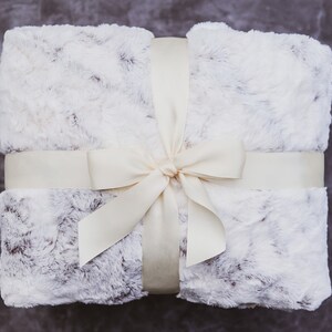May include: A white faux fur throw blanket tied with a cream-colored satin ribbon and bow.