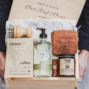 Puede incluir: Una caja de madera llena de regalos de inauguraci&oacute;n de la casa, que incluyen una botella de jab&oacute;n para lavar platos, una bolsa de caf&eacute;, una vela y un posavasos de madera con el texto "HOME SWEET HOME Chris & Diana 2023".
