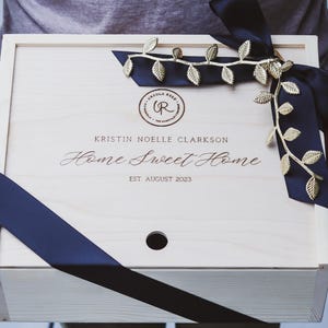 Puede incluir: Una caja de madera con una cinta negra y detalles de hojas doradas. La caja est&aacute; grabada con el texto "Kristin Noelle Clarkson Home Sweet Home Est August 2023".