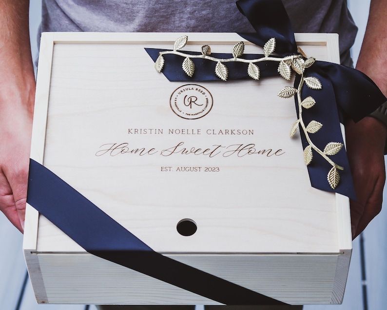 Puede incluir: Una caja de madera con una cinta negra y detalles de hojas doradas. La caja est&aacute; grabada con el texto "Kristin Noelle Clarkson Home Sweet Home Est August 2023".