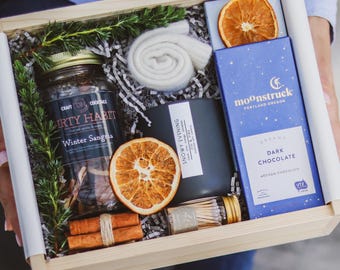 May include: A wooden gift box filled with winter-themed items, including a jar of sangria, a candle, a bar of dark chocolate, a pair of cinnamon sticks, a dried orange slice, and a white knitted scarf. The box is labeled "moonstruck" and "Portland Oregon".