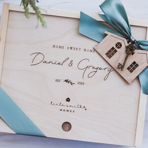 Puede incluir: Una caja de madera con una cinta azul y un recorte de casa de madera con una llave. La caja est&aacute; grabada con el texto "Home Sweet Home", "Daniel & Gregory", "Est. 2023", y "Lila Smiths Homes".