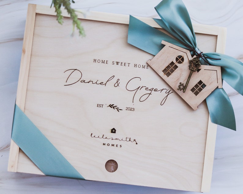Puede incluir: Una caja de madera con una cinta azul y un recorte de casa de madera con una llave. La caja est&aacute; grabada con el texto "Home Sweet Home", "Daniel & Gregory", "Est. 2023", y "Lila Smiths Homes".