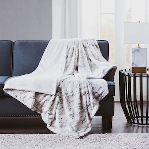 May include: A white and gray faux fur throw blanket draped over a dark gray couch. The blanket has a soft, fluffy texture and is perfect for adding warmth and comfort to any room.