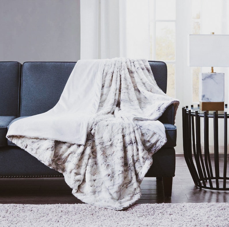 May include: A white and gray faux fur throw blanket draped over a dark gray couch. The blanket has a soft, fluffy texture and is perfect for adding warmth and comfort to any room.