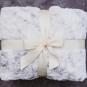 May include: A white faux fur throw blanket tied with a cream-colored satin ribbon and bow.