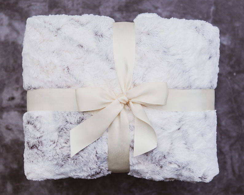 May include: A white faux fur throw blanket tied with a cream-colored satin ribbon and bow.