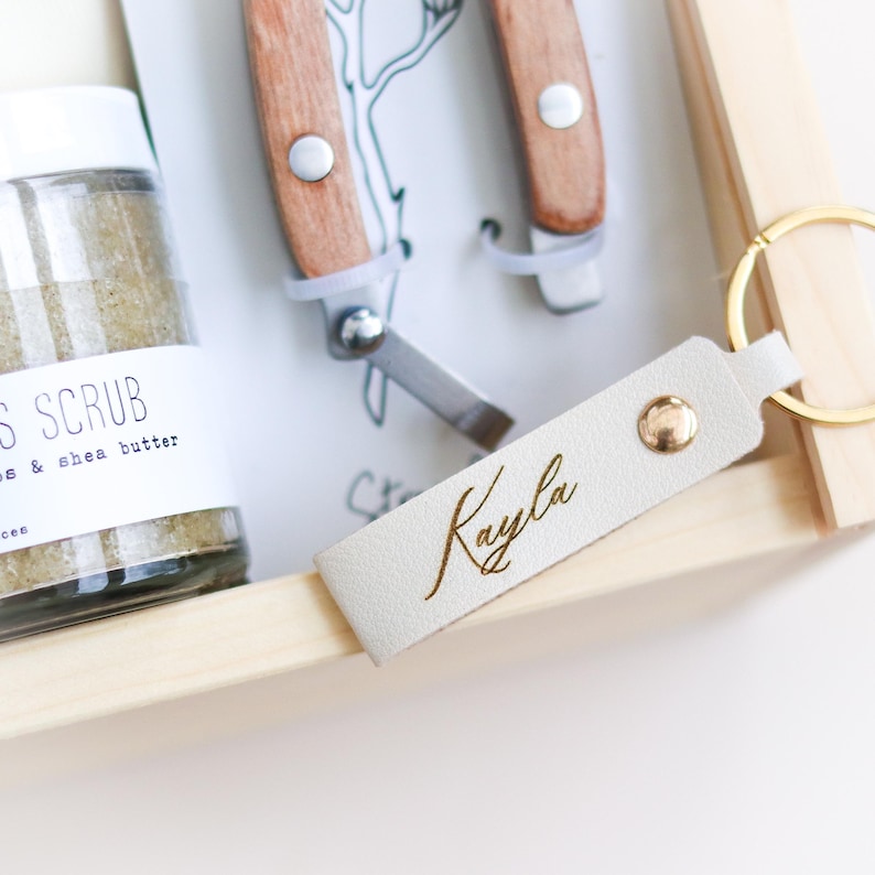 May include: A white leather keychain with the name "Kayla" engraved in gold lettering. The keychain is attached to a gold key ring. The keychain is in a wooden box with a jar of body scrub and two small tools.