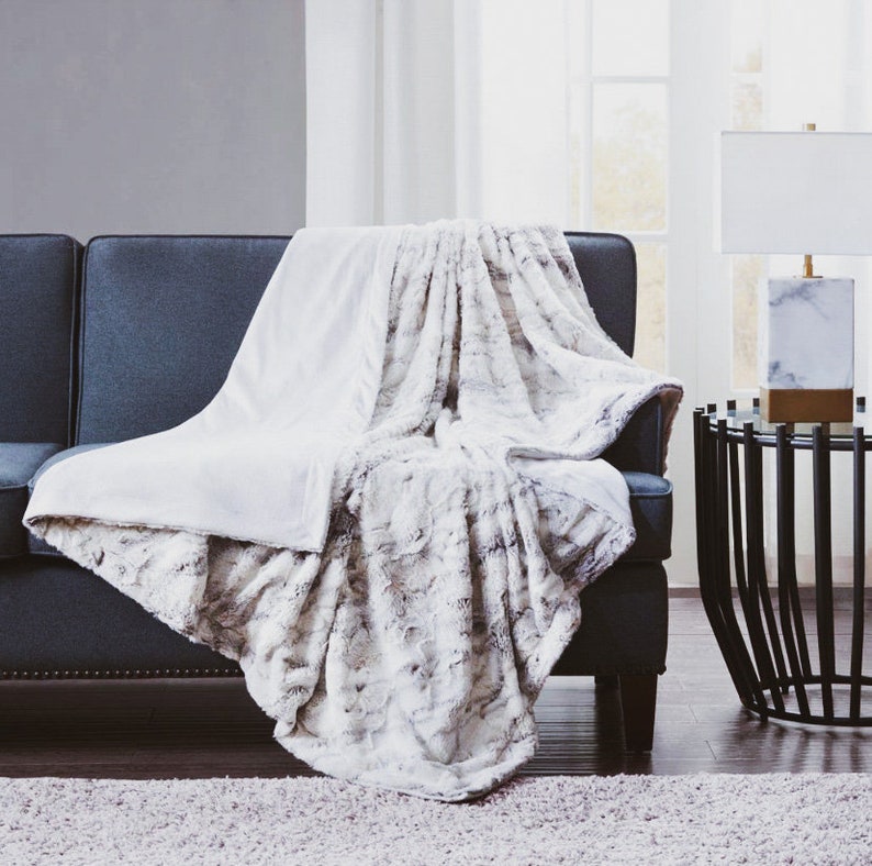 Puede incluir: Una manta de piel sint&eacute;tica blanca y gris drapeada sobre un sof&aacute; gris oscuro. La manta tiene una textura suave y esponjosa y es perfecta para agregar calidez y comodidad a cualquier habitaci&oacute;n.
