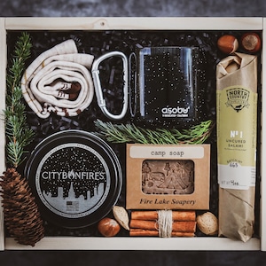 May include: A wooden gift box filled with camping and outdoor themed items. The box contains a black and white candle, a bar of soap, a black insulated mug, a brown paper wrapped salami, a pair of socks, and cinnamon sticks.