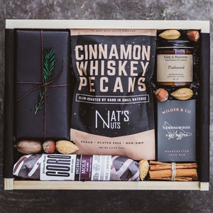May include: A wooden gift box filled with a variety of gourmet treats, including cinnamon whiskey pecans, a teakwood scented candle, a sandalwood and patchouli soap bar, and a salami. The box is wrapped in black paper with a sprig of rosemary.