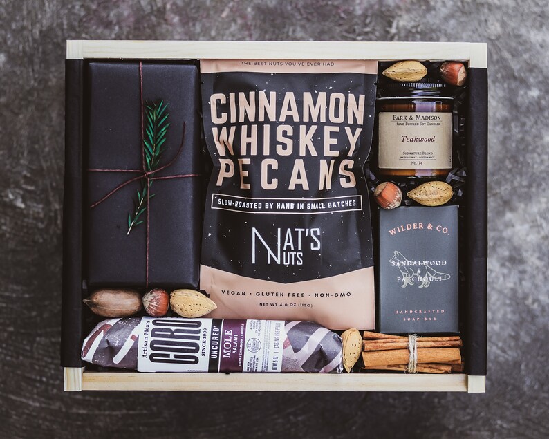 May include: A wooden gift box filled with a variety of gourmet treats, including cinnamon whiskey pecans, a teakwood scented candle, a sandalwood and patchouli soap bar, and a salami. The box is wrapped in black paper with a sprig of rosemary.