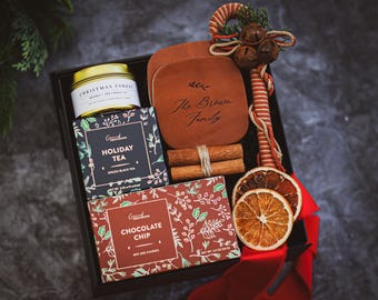 May include: A gift box with a candle, tea, and chocolate chip cookies. The box includes a wooden coaster with "The Brown Family" engraved on it, cinnamon sticks, and dried orange slices. A decorative candy cane and red ribbon complete the holiday-themed gift.