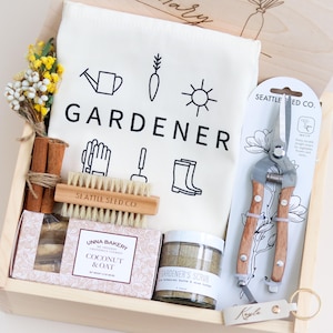 May include: A wooden gift box filled with gardening themed items. The box contains a white tea towel with the word "GARDENER" printed on it, a pair of gardening shears, a small brush, a bar of soap, a jar of scrub, and a package of cookies. The box is decorated with a small wooden tag with the name "Kayle" written on it.