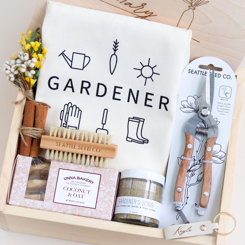 May include: A wooden gift box filled with gardening themed items. The box contains a white tea towel with the word "GARDENER" printed on it, a pair of gardening shears, a small brush, a bar of soap, a jar of scrub, and a package of cookies. The box is decorated with a small wooden tag with the name "Kayle" written on it.
