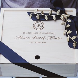 May include: A wooden box with a black ribbon and gold leaf accents. The box is engraved with the text "Kristin Noelle Clarkson Home Sweet Home Est August 2023".