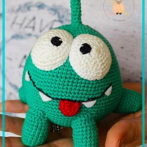 May include: Green crocheted alien plush toy with large white eyes and a red tongue sticking out. The toy has a long green antenna on top of its head and four short legs.