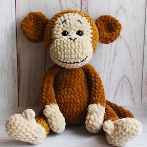 May include: A crocheted monkey plush toy with brown fur and a cream-coloured face. The monkey is sitting with its legs crossed and has a friendly expression.