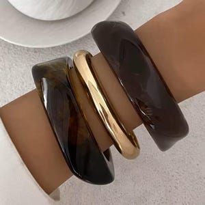May include: A collection of four bracelets. Two are a dark brown, translucent material, one is a solid gold color, and the fourth is a lighter brown, translucent material. The bracelets are stacked on a wrist.