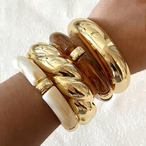 May include: A stack of four bracelets featuring a smooth gold-tone bangle, a dark brown translucent resin bangle, a gold-tone twisted rope-style bangle, and a white marbled resin bangle with a gold-tone metal accent piece.