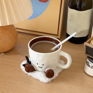 May include: A white ceramic mug with a brown interior, filled with coffee and a white straw. The mug has a smiling face design and sits on a fluffy white coaster with brown accents. A charming and whimsical coffee cup.
