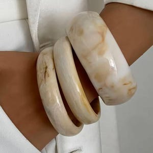 Puede incluir: Tres pulseras de color marfil con efecto veteado. Las pulseras tienen un acabado brillante y anchos variables. La paleta de colores incluye tonos crema, beige y marrón claro. Estos accesorios son adecuados para el uso diario.