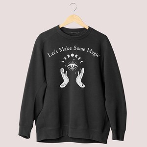 May include: Black sweatshirt with a white graphic design featuring a pair of hands holding a crescent moon and stars with an eye in the center. The text "Let's Make Some Magic" is printed above the graphic.