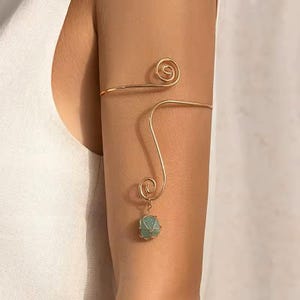 Op de afbeelding: Een goudkleurige metalen armband voor de bovenarm met een glad, gebogen draadontwerp en twee spiraalvormige uiteinden. Eén uiteinde van de draad loopt naar beneden en houdt een kleine, lichtgroene, ruwe steen hanger in een fijn gouden draadmandje.