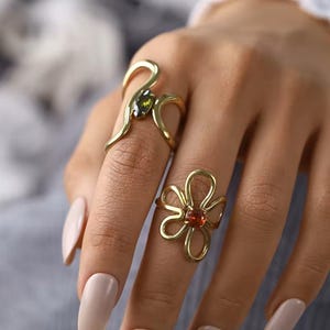 May include: Two gold-colored rings. One ring features a curved design with a green gemstone. The other ring is shaped like a flower with a red gemstone in the center. The rings are on a hand with light pink nail polish.