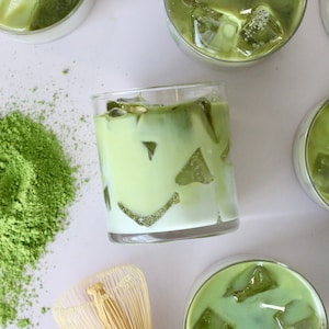 May include: Overhead shot of several glasses of iced matcha latte with ice cubes, arranged on a white surface. A pile of matcha powder and a bamboo whisk are also visible, suggesting the preparation of the beverage.