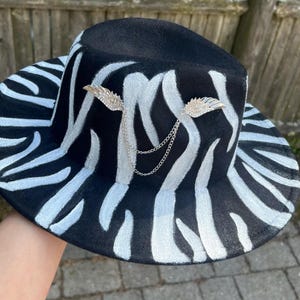May include: Black fedora hat with a zebra stripe pattern in white. The hat features silver-colored wing accents connected by a delicate chain. The hat has a wide brim and a classic fedora shape.