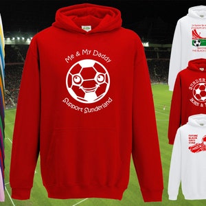 Puede incluir: Sudadera con capucha roja con un gráfico de balón de fútbol blanco y el texto "Me & My Daddy Support Sunderland". La sudadera se muestra sobre un fondo de hierba verde.