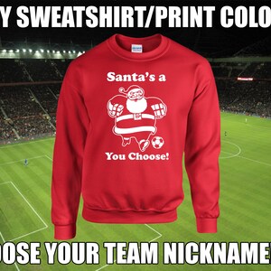 Pode incluir: Camisola vermelha com um gráfico branco do Papai Noel jogando futebol. O texto "Santa's a You Choose!" está impresso na camisola.