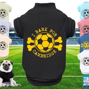 May include: A black dog shirt with the text "I Bark for Cambridge" and a yellow soccer ball design. Other shirts in the image are light blue, pink, white, and beige, each with a soccer ball and the text "Cambridge Born & Bred" or "Cambridge Supporter". A pug dog is wearing a black shirt.